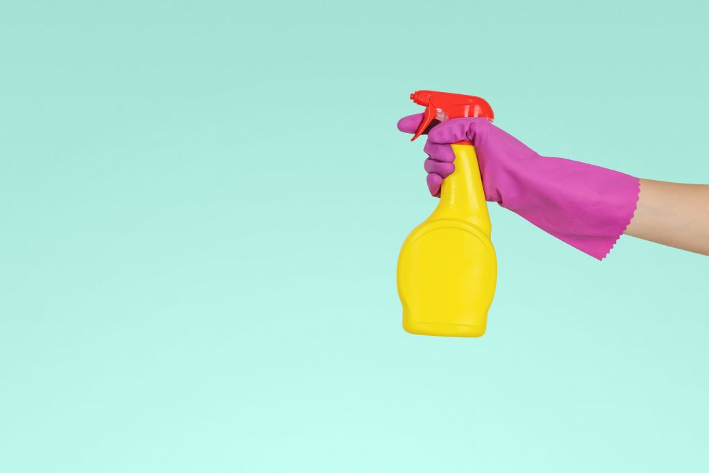 Hand in einem lila Handschuh hält eine gelbe Sprühflasche vor türkisem Hintergrund